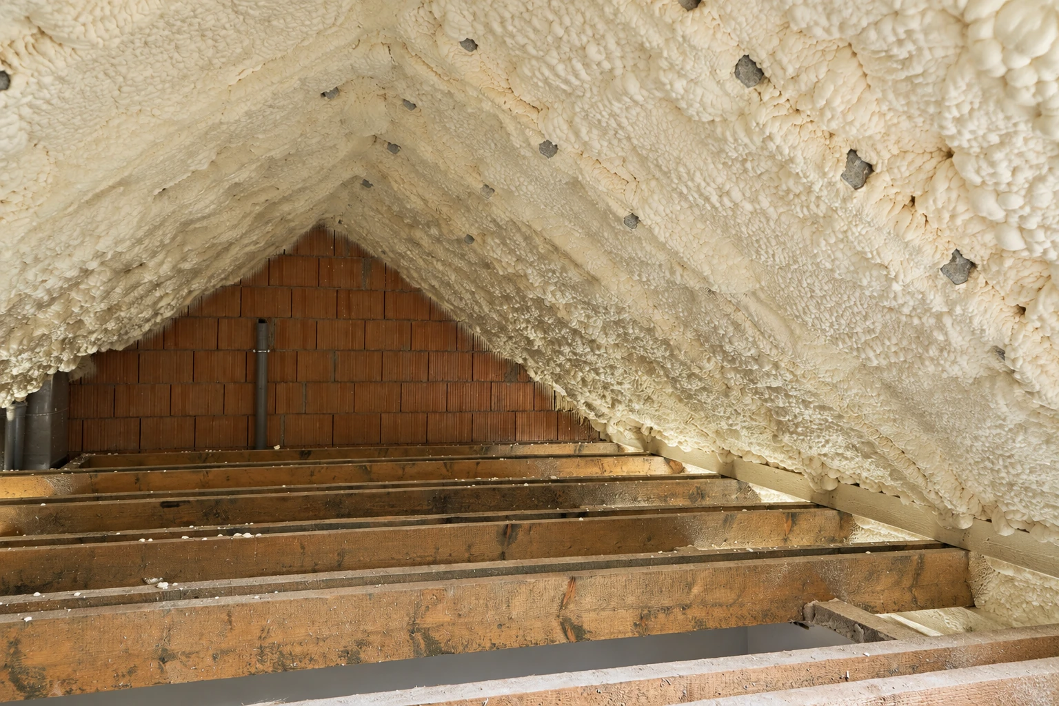 Spray foam coats the sloped roof of an attic above exposed wooden joists and a brick gable wall.
