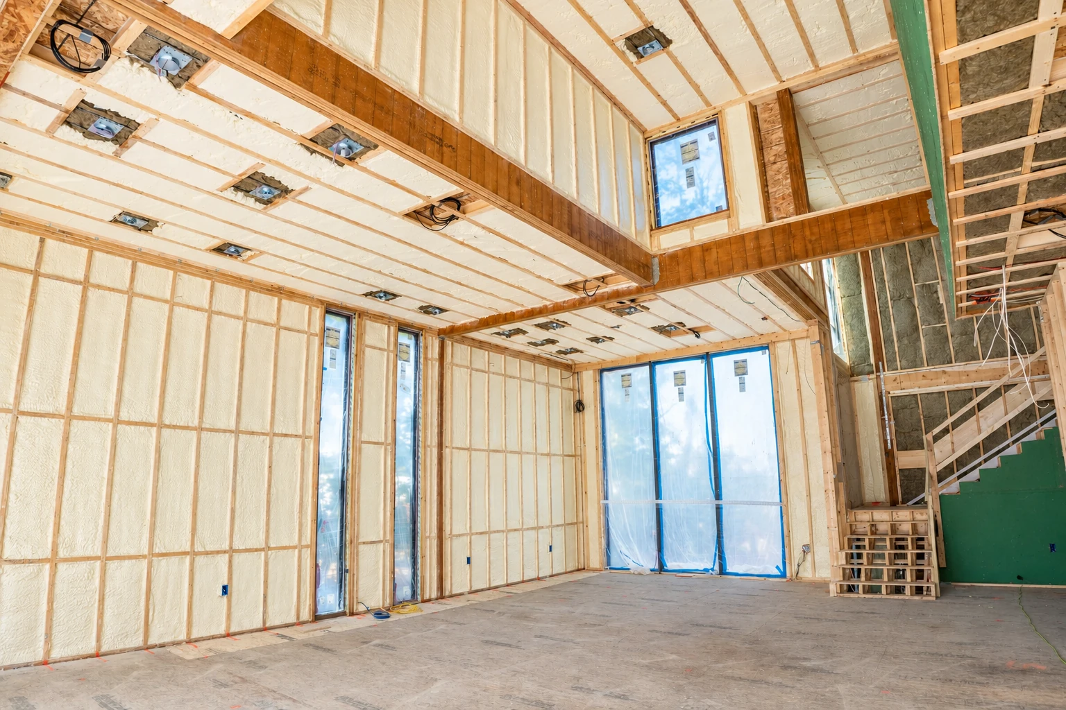 An unfinished room has spray foam insulation between wall studs and roof framing, with windows covered in protective film.
