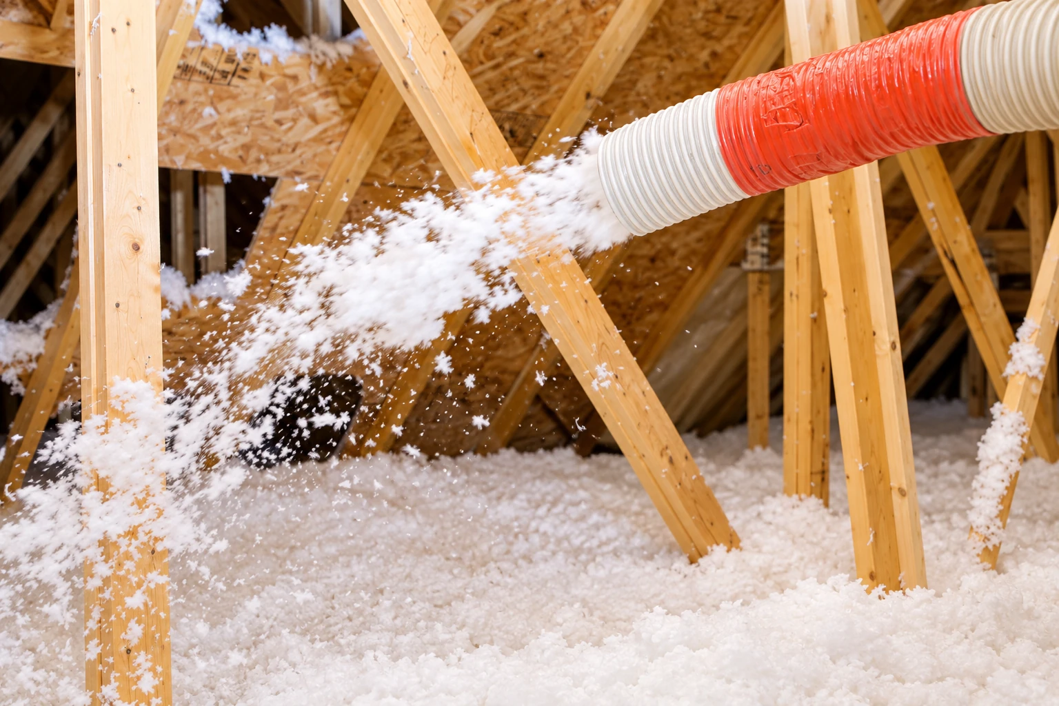 Blown-in insulation sprays from a red-and-white hose across the floor of a wooden attic.