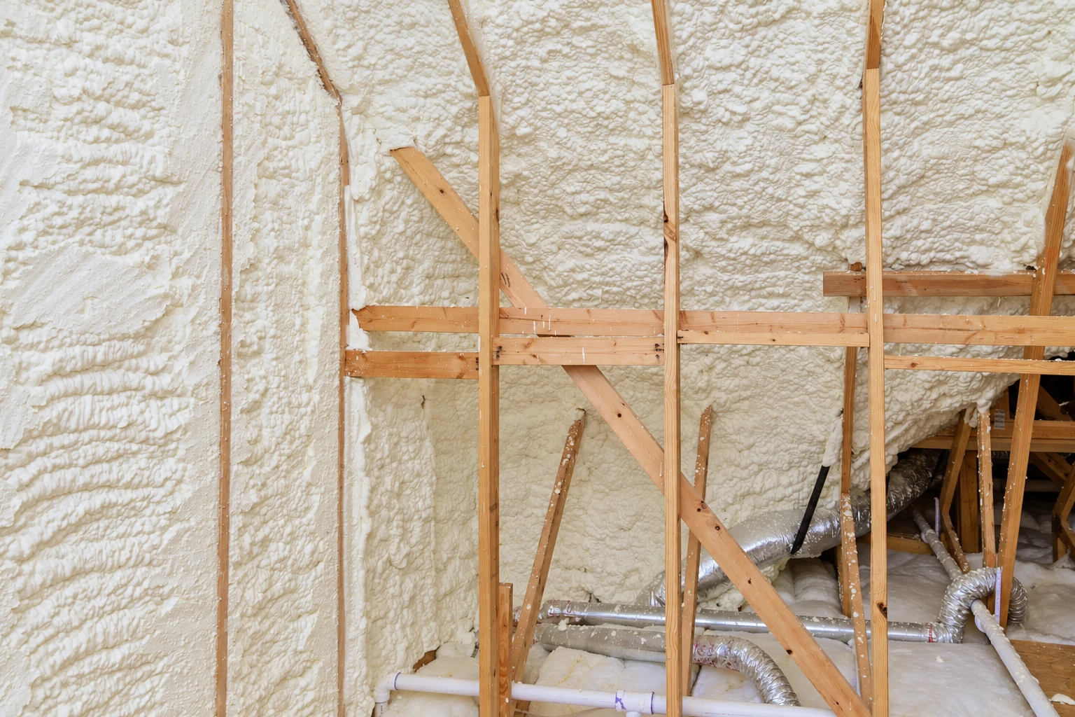 Open wall framing and roof rafters are covered with thick white spray foam insulation, with ducts and pipes below.